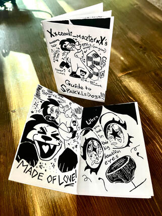 Display photo of a black and white mini zine. Cover: An annotated drawing of a sparkle dog. Open spread: The same dog excitedly speaking to the viewer. The text reads: "Sparkledogz R made of love! When you look in their big beautiful eyes..."
